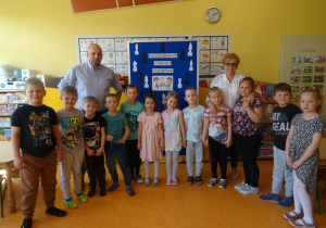 Pani dyrektor Maria Królikowska i pan Szadkowski stoją pośród dzieci, w tle tablica z napisem przedszkolny turniej szachowy.